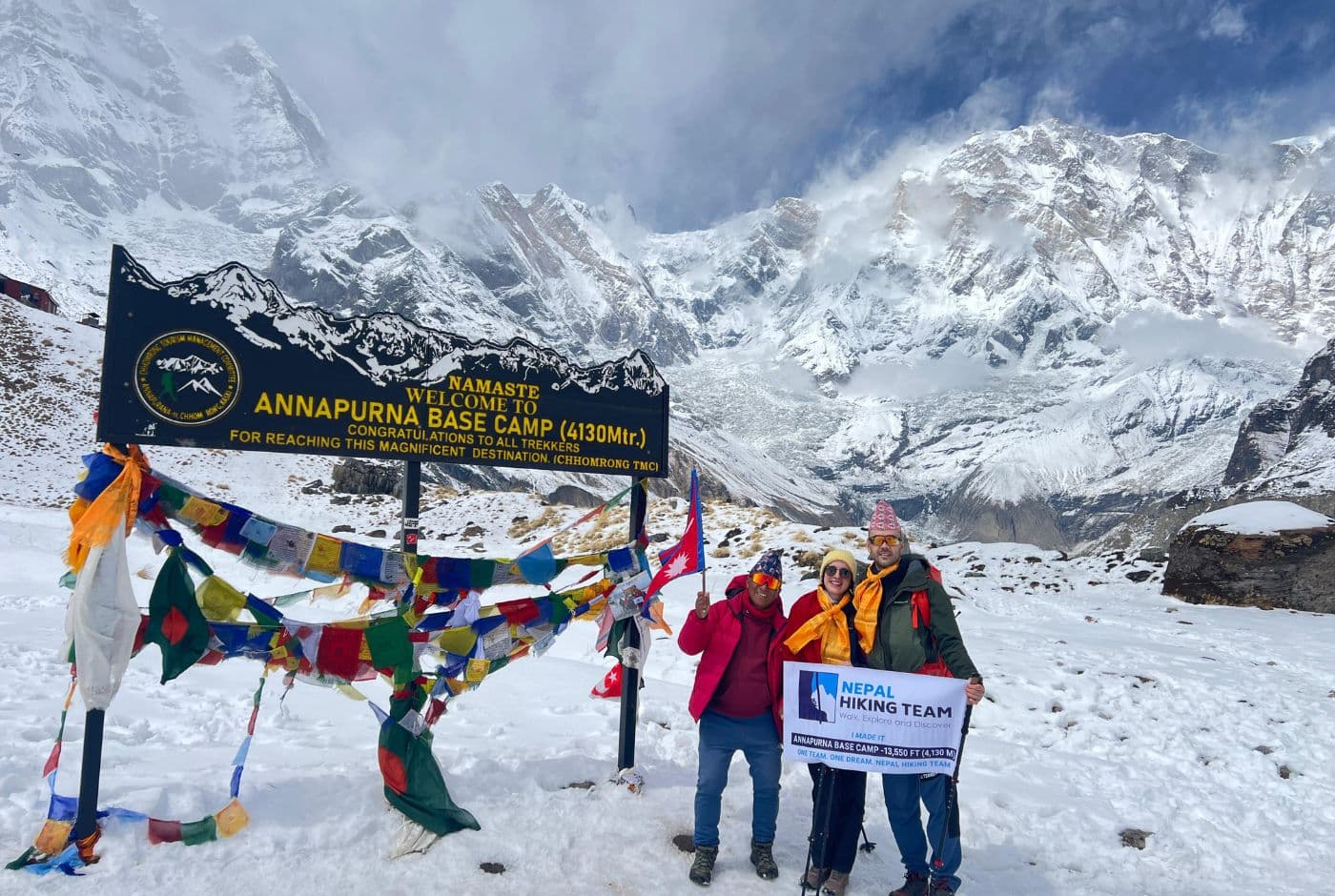 Trekkers celebrating at Annapurna Base Camp (4,130m) with Nepal Hiking Team banner, prayer flags, and snow-covered Annapurna peaks in the background