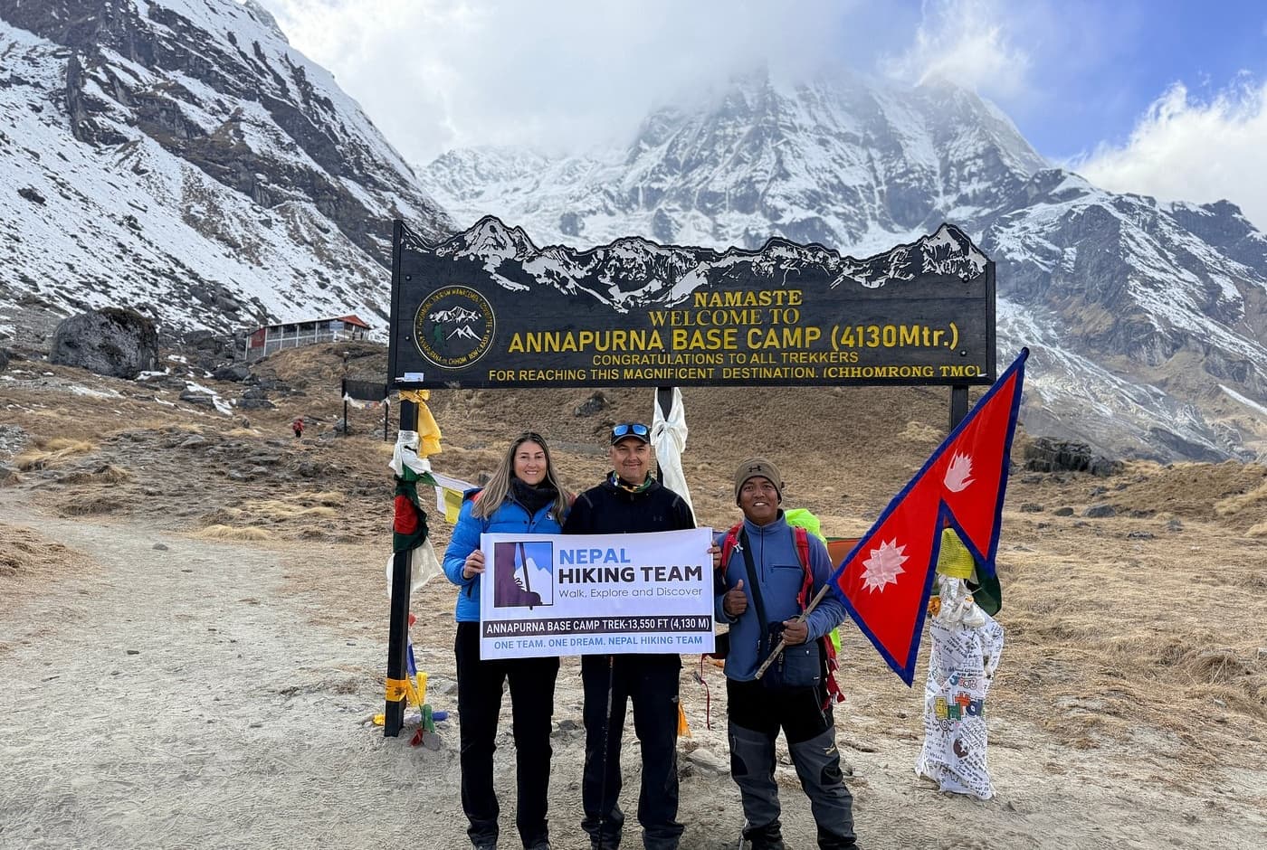 Trekkers with Nepal Hiking Team holding a banner and Nepal flag at Annapurna Base Camp (4,130m), with Annapurna massif and cloudy peaks behind