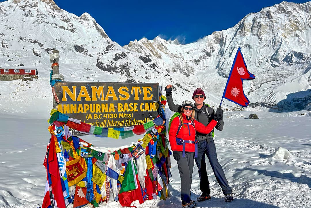 Trekkers celebrating at Annapurna Base Camp signboard at 4130 meters with Nepali flag, prayer flags, and snow-covered Annapurna massif peaks in background