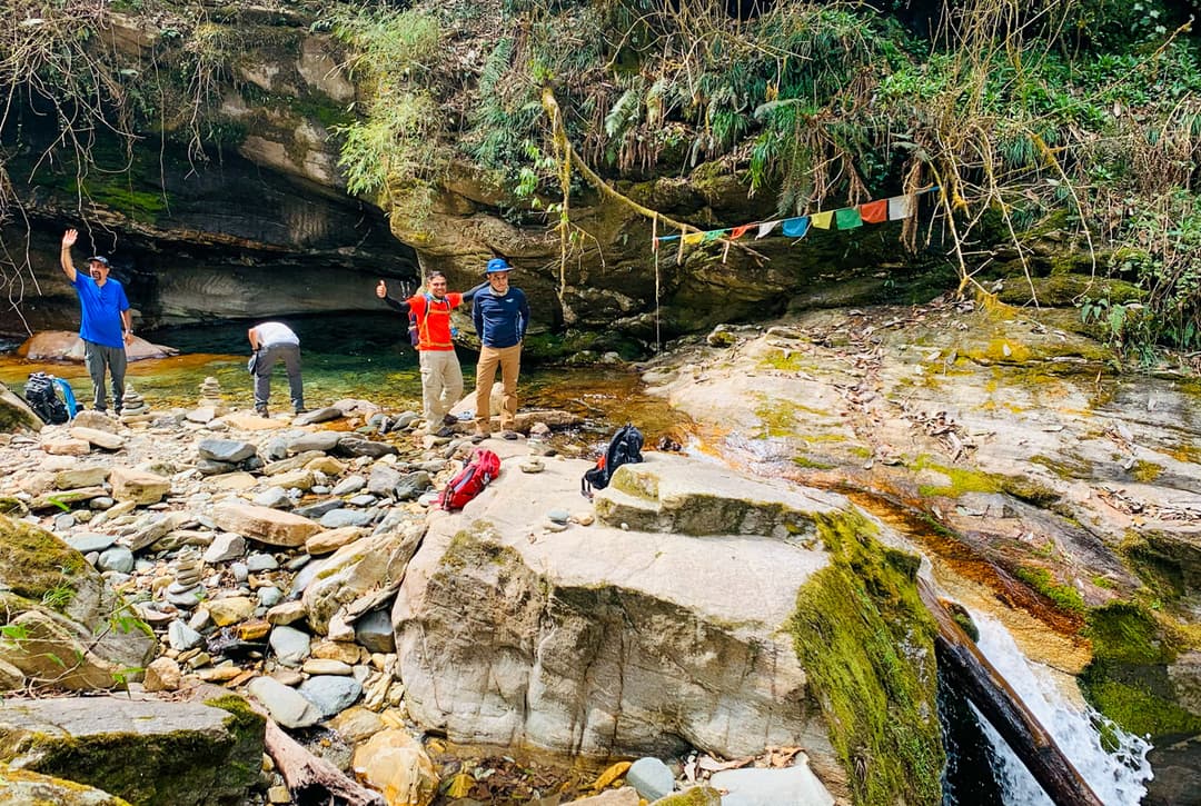 Trekkers Resting At Mossy Stone Water Source With Prayer Flags In Rhododendron Forest On Annapurna Base Camp Trek Route To Ghorepani